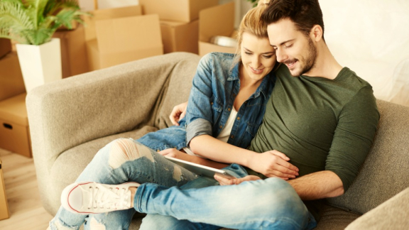 Ein glückliches Paar sitzt entspannt auf dem Sofa zwischen Umzugskartons und berechnet den USA-Umzug mit dem Online-Rechner von DACHSER & KOLB auf einem Tablet.