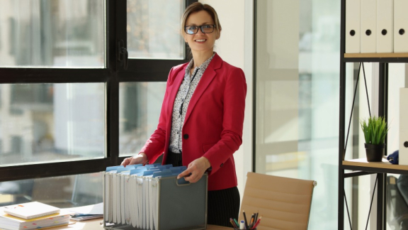 Eine lächelnde Geschäftsfrau im roten Blazer sortiert Aktenordner in einer Archivbox im Büro, passend zum Kleintransport für Geschäftskunden mit DACHSER & KOLB.