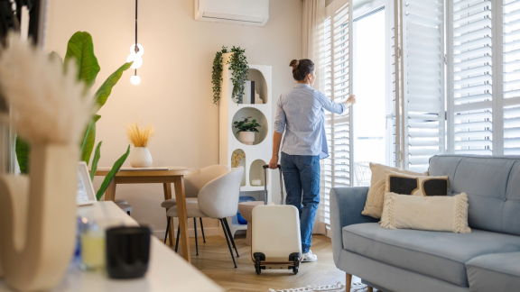 Frau im neuen Zuhause, öffnet Jalousien und zieht einen Koffer hinter sich her. Symbol für Relocation Service für Expats.