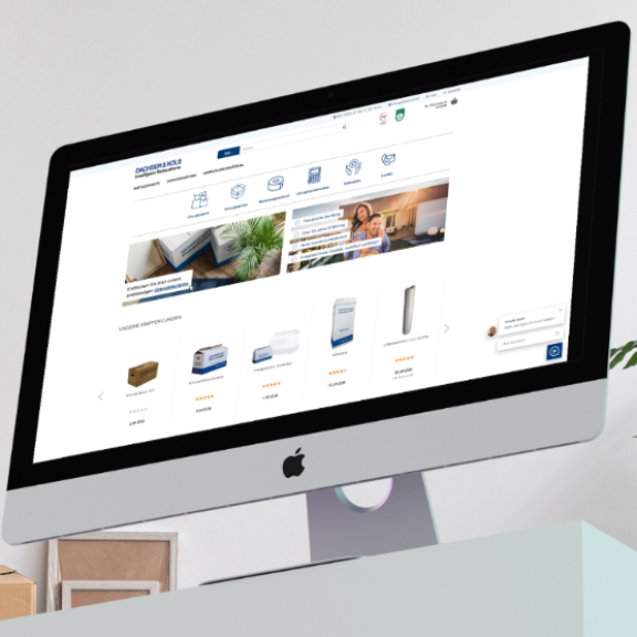 Ein Schreibtisch mit PC zeigt den Umzugsshop DACHSER & KOLB, umgeben von Umzugskartons und Pflanzen in einem Büro.