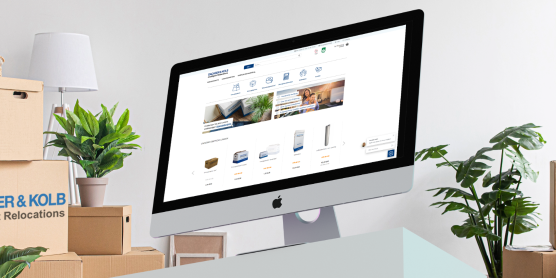 Ein Schreibtisch mit PC zeigt den Umzugsshop DACHSER & KOLB, umgeben von Umzugskartons und Pflanzen in einem Büro.