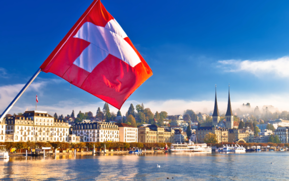 Schweizer Flagge am See mit Stadtansicht – Symbolbild für einen Umzug von Deutschland in die Schweiz
