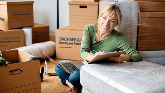 Frau lächelt mit Umzugsplanung auf Klemmbrett zwischen Umzugskartons.