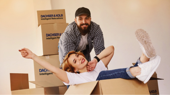 Lächelndes Paar beim Umzug, Frau sitzt in DACHSER & KOLB Umzugskarton – Qualität und Zuverlässigkeit durch FIDI-Zertifizierung.