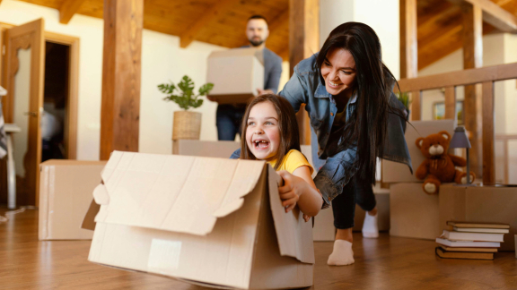 Glückliche Familie beim Umzug. Mädchen lacht in Umzugskarton, Mutter schiebt, Vater trägt Kartons. Umzug auf Ratenzahlung.