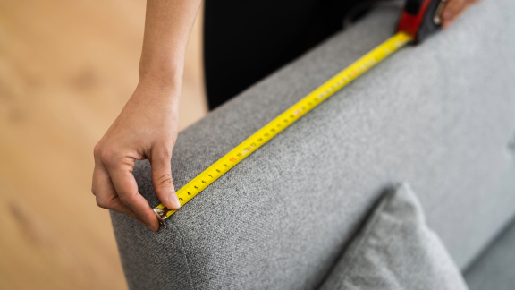 Eine Person misst mit einem Maßband die Maße eines Sofas, um das Volumengewicht für den Transport zu ermitteln und die Umzugsplanung zu erleichtern.