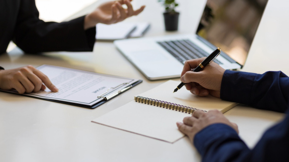 Zwei Personen an einem Schreibtisch: Eine macht sich Notizen, die andere zeigt auf Unterlagen. Symbol für Relocation Service für Privatpersonen.