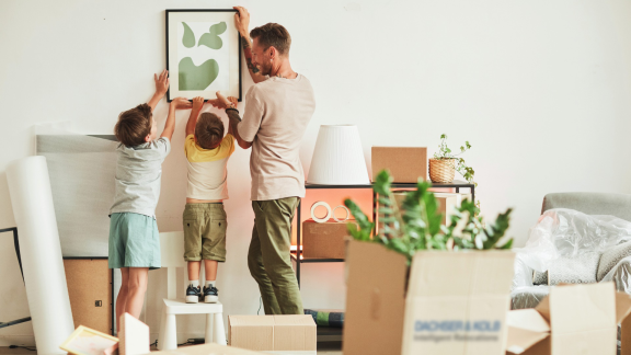 Familie hängt Bild in neuer Wohnung auf, Umzugskartons im Raum – Günstig umziehen