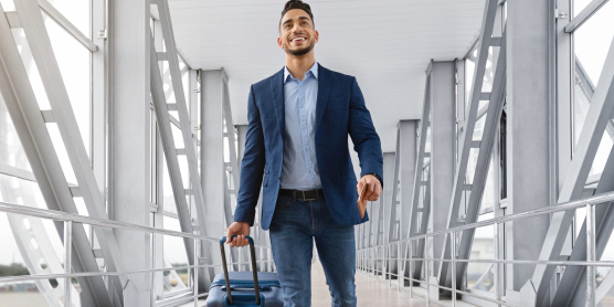 Ein lächelnder Expat im Business-Outfit zieht seinen Rollkoffer durch ein modernes Flughafenterminal zu einem internationalen Auslandseinsatz.