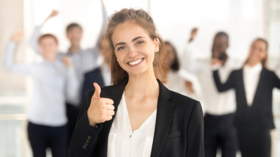 Eine lächelnde HR-Managerin zeigt den Daumen nach oben mit einem jubelnden Business-Team im Hintergrund nach einem erfolgreichen Relocation-Prozess.