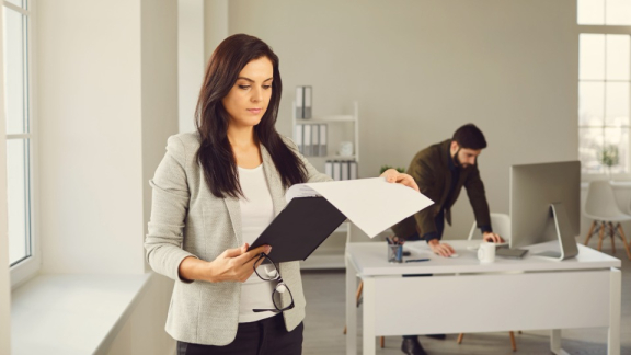 Eine Facility Managerin prüft Dokumente zur Büro-Umstrukturierung im hellen Office, im Hintergrund ein Kollege am Schreibtisch – Gebäudelogistik für Facility Management.